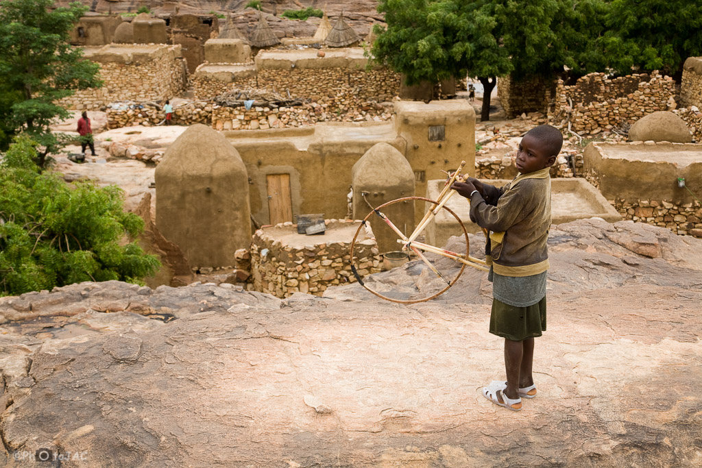 País Dogón. Mali. Un niño con un juguete (una rueda) totalmente artesanal. Al fondo viviendas y graneros erigidos con madera, adobe y piedra en la aldea de Daga.