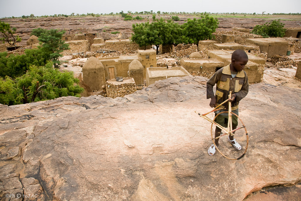 País Dogón. Mali. Un niño con un juguete (una rueda) totalmente artesanal. Al fondo viviendas y graneros erigidos con madera, adobe y piedra en la aldea de Daga.