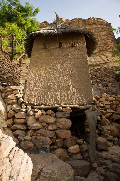 País Dogón. Mali. Aldea de Nombori. Granero erigido con madera y adobe y soportado por piedras y un pilar de madera. Al fondo las paredes de la falla de Bandiagara.